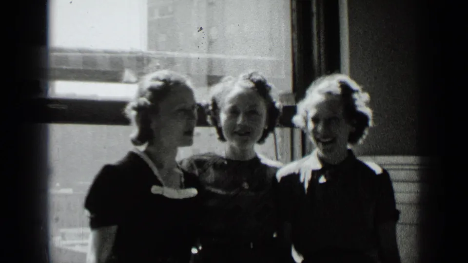 Three young women in 1930s attire stand close together by a bright window in a New York interior.