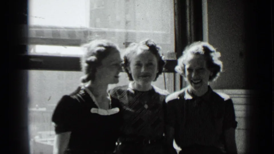 Three young women in 1930s dress stand shoulder to shoulder in a windowed New York interior.