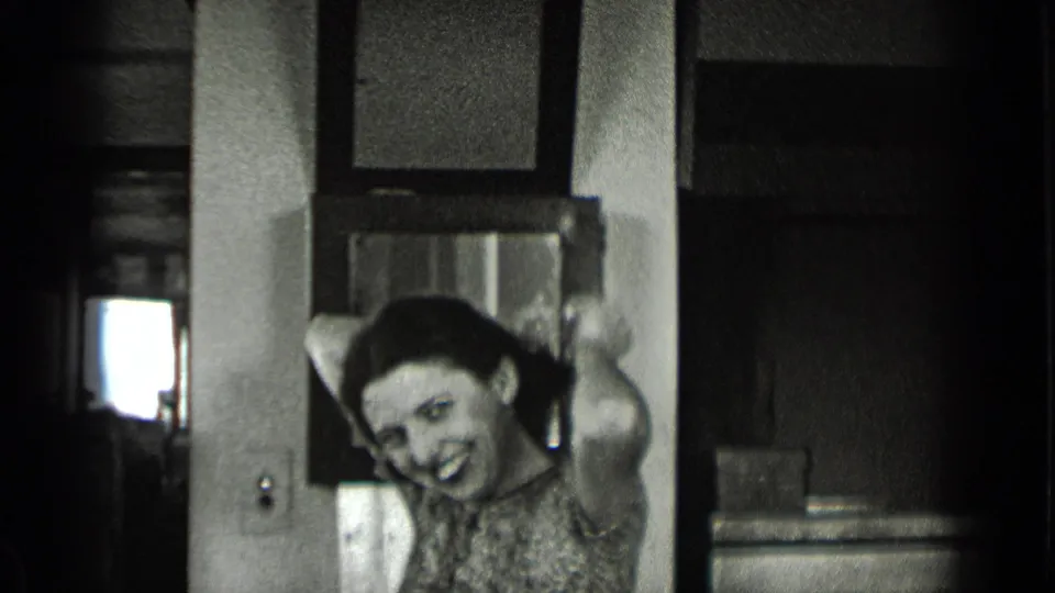 A woman in a patterned dress smiles from behind an interior doorway in 1939 New York footage, hand raised near her bow or hair.