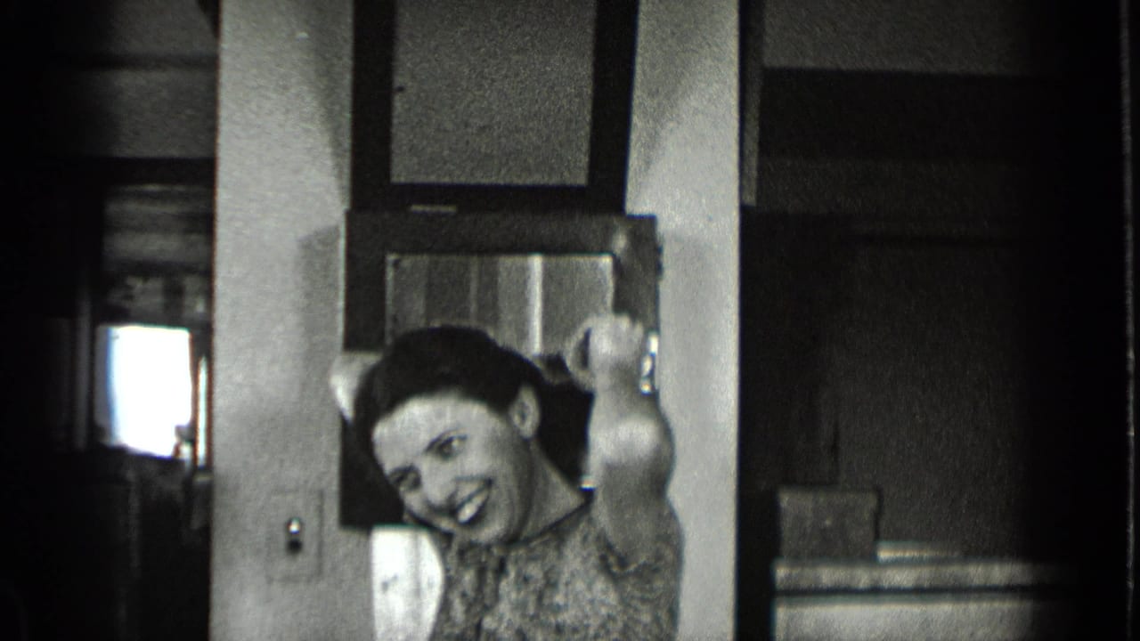 In a 1939 New York office interior, a woman in a patterned dress smiles toward the camera with one hand raised near a desk.