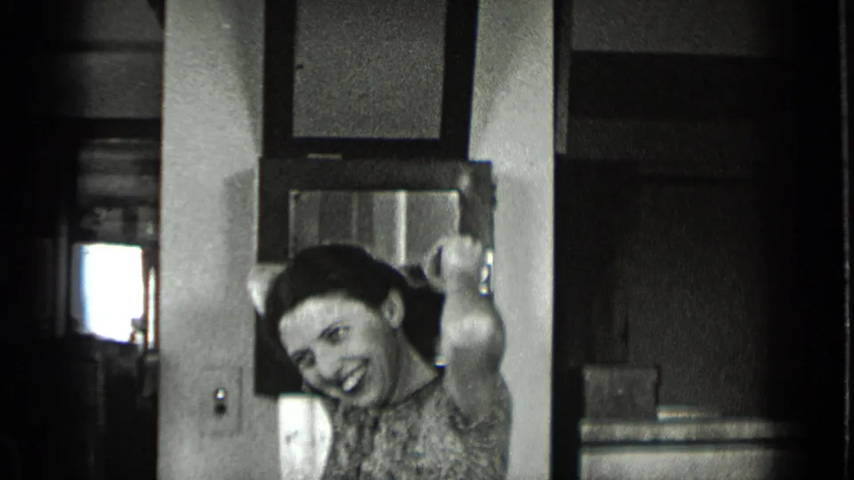 In a 1939 New York office interior, a woman in a patterned dress smiles toward the camera with one hand raised near a desk.