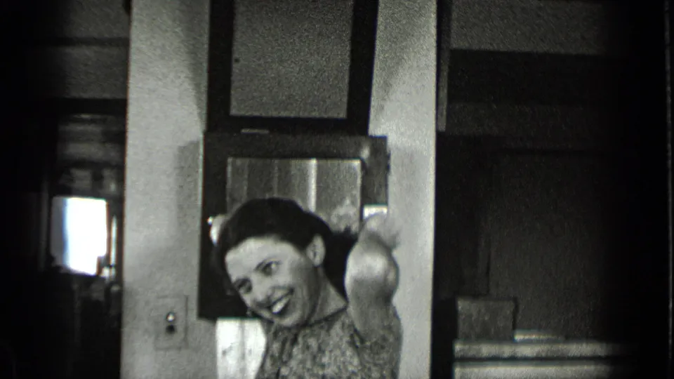 A woman in a patterned dress smiles in a 1939 New York office, framed by a desk, wall surfaces, and a dark doorway behind her.