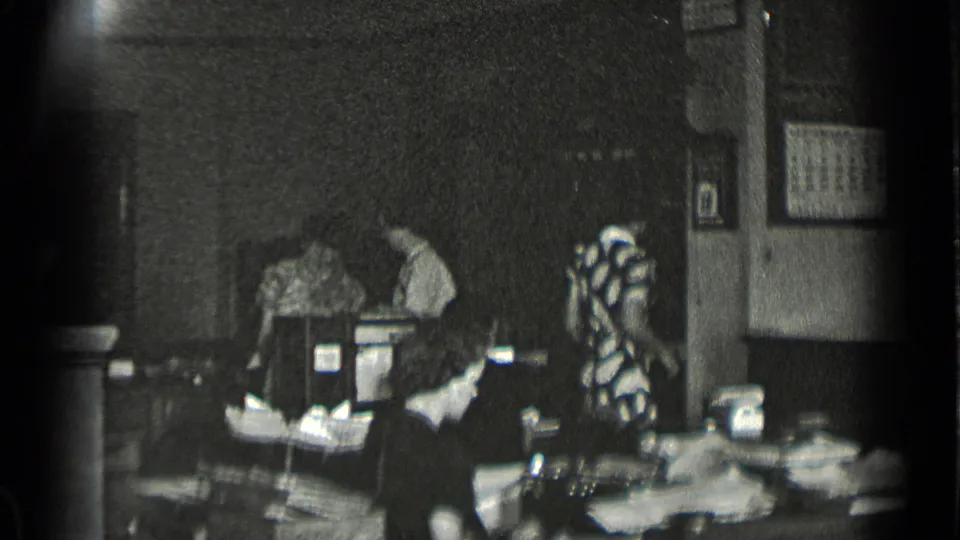 Workers sit at desk stations in a dim 1939 New York office interior, with papers, wall frames, and office equipment visible.