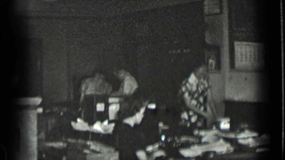 1939 New York home-movie frame showing women at worktables with fabric in a dark sewing or garment-room interior.