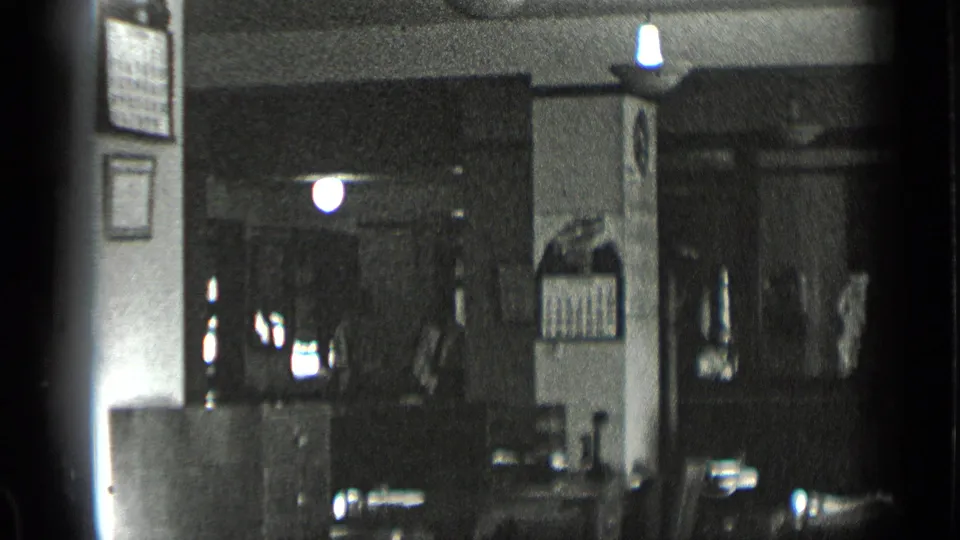 1939 New York 8mm frame showing the sewing shop interior with counters, notices, overhead lights, and shadowed work areas.