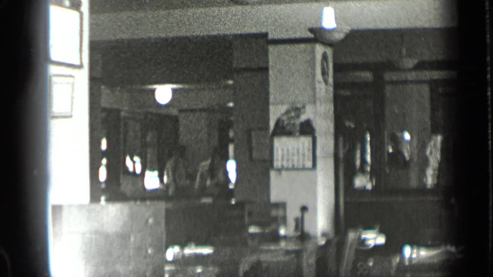 1939 New York home-movie view across a sewing shop interior, with a bright side wall, hanging light, counter, and shadowed rear workspace.