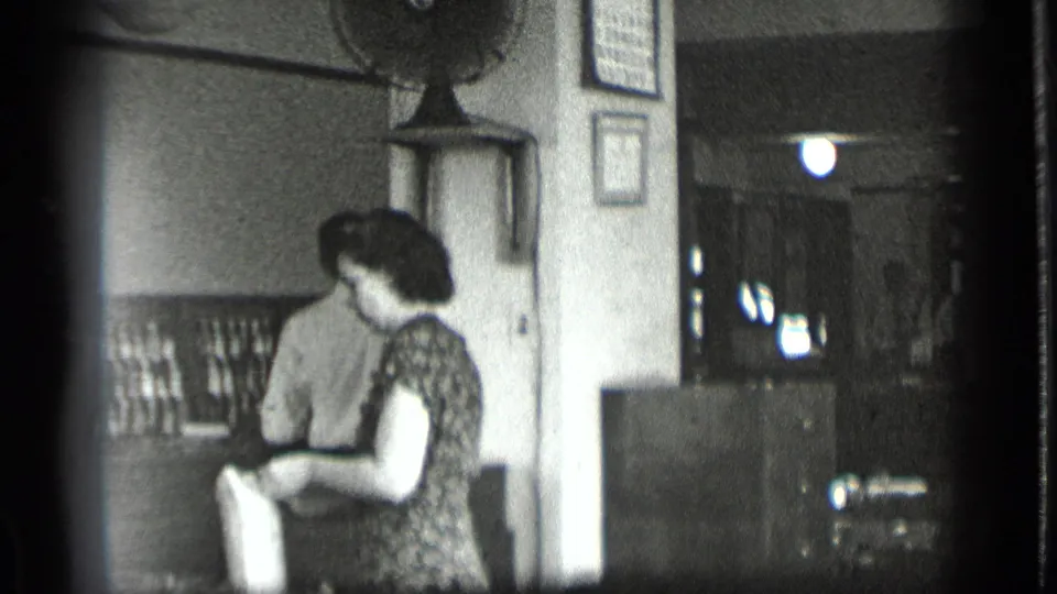 1939 New York frame showing a woman in a patterned dress holding fabric inside the sewing shop, with wall notices and cabinets nearby.