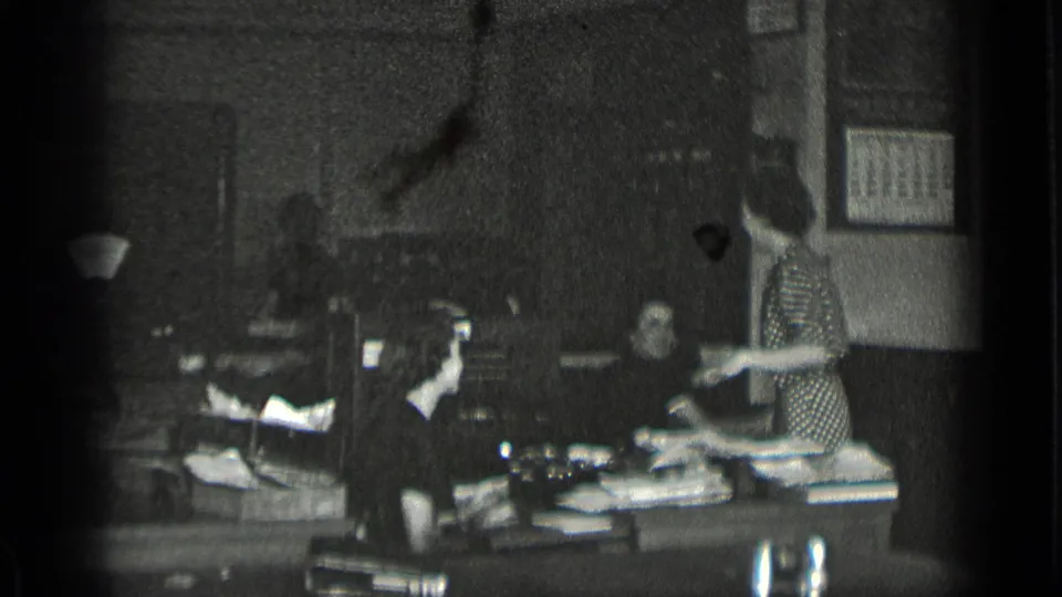 Black-and-white 1939 New York office frame with women handling papers near a desk, wall calendar, and period office furnishings.