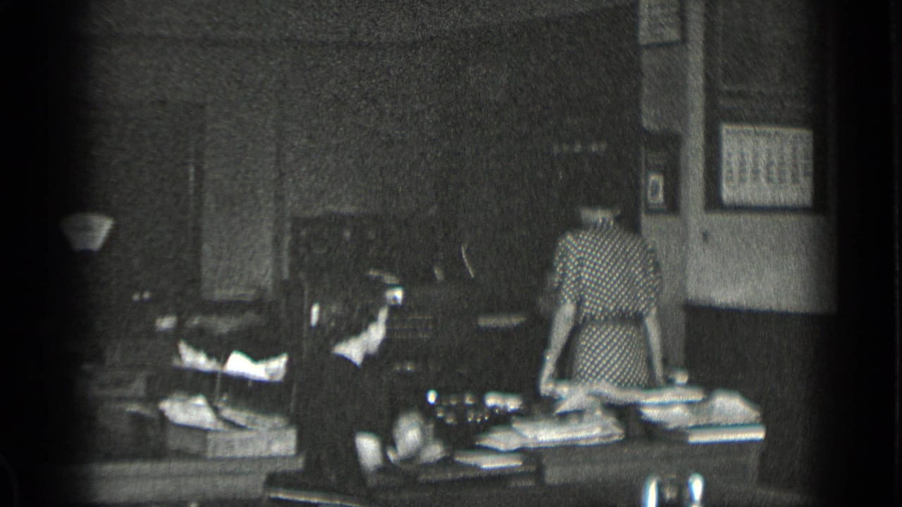 Woman in a patterned dress stands beside a desk while another woman sits nearby.