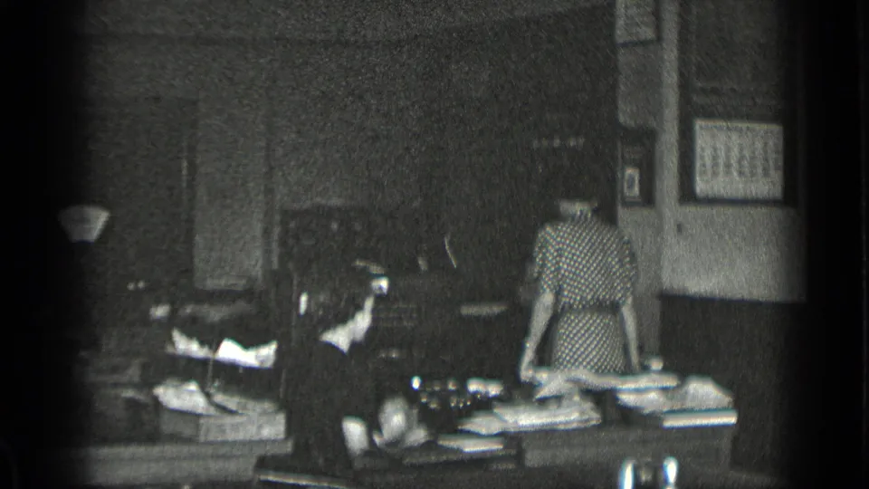 Black-and-white 1939 New York office view with a woman in a patterned dress standing at a paper-covered desk and another seated worker nearby.