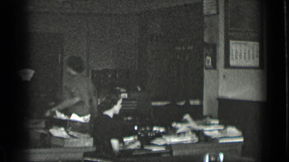 Black-and-white 1939 New York office frame showing a seated worker at a paper-strewn desk with motion blur from another person nearby.