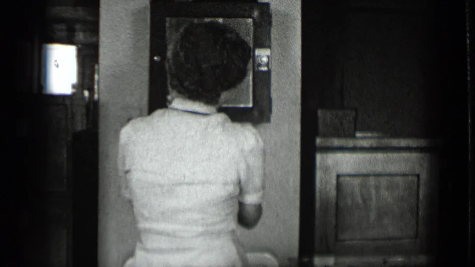 Black-and-white 1939 New York office frame with a woman in a light uniform facing a wall-mounted device beside a dark cabinet or partition.