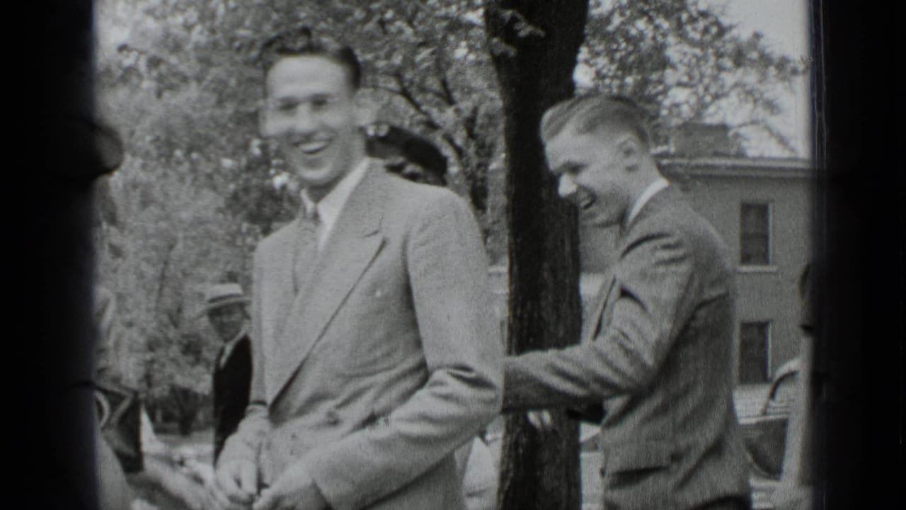 Two young men in suits stand smiling outdoors near trees and a building.