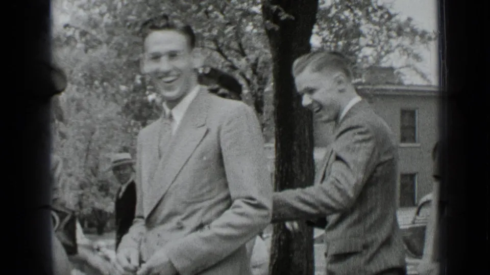 1939 New York home-movie frame of young men in suits smiling and joking outdoors near trees and an older building.