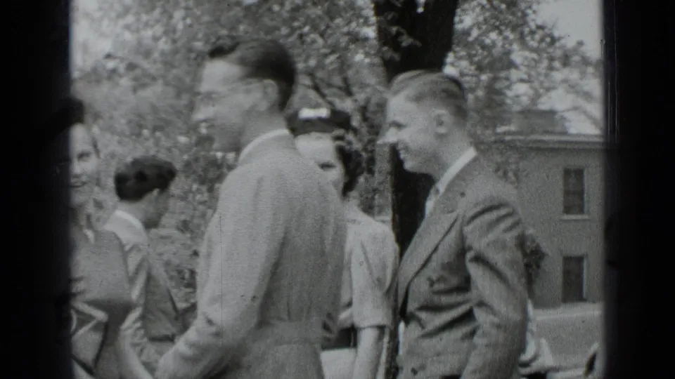 Young adults in suits and dresses gather in a New York outdoor setting, with leafy trees and an older building visible behind them.