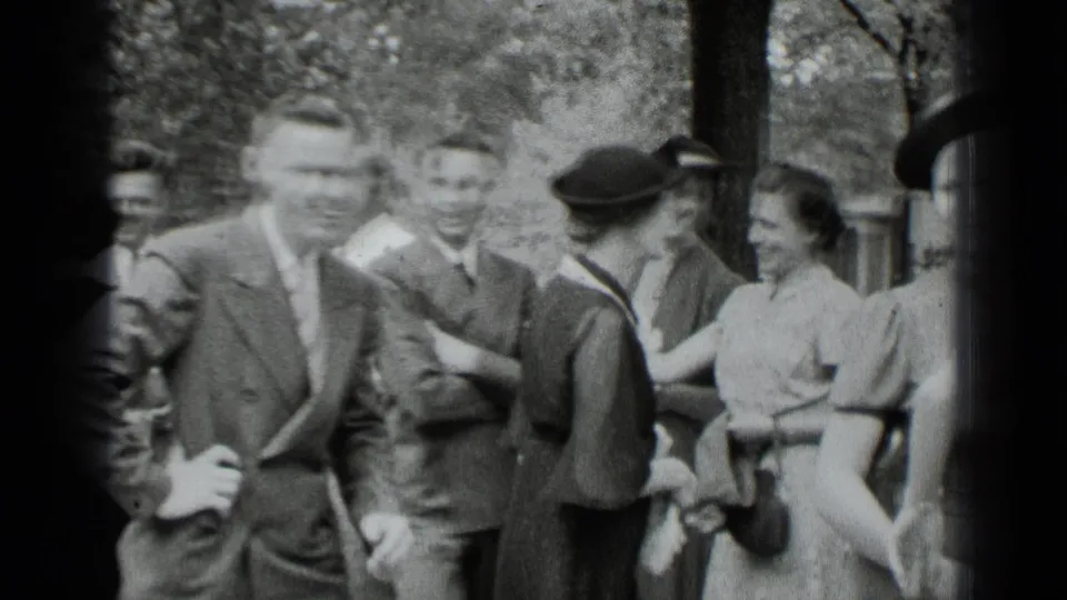 In 1939 New York home-movie footage, adults in suits, hats, and dresses gather and greet each other near trees.
