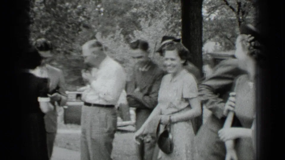 A woman in a light dress smiles toward the camera while men in shirts and jackets gather nearby in New York.
