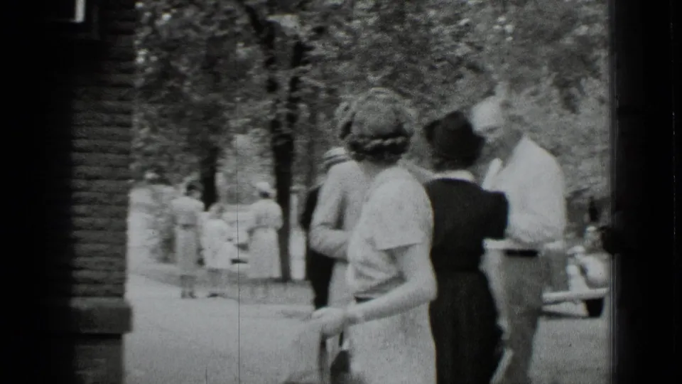 A woman in a short-sleeved dress walks through a shaded New York park area as men stand nearby.
