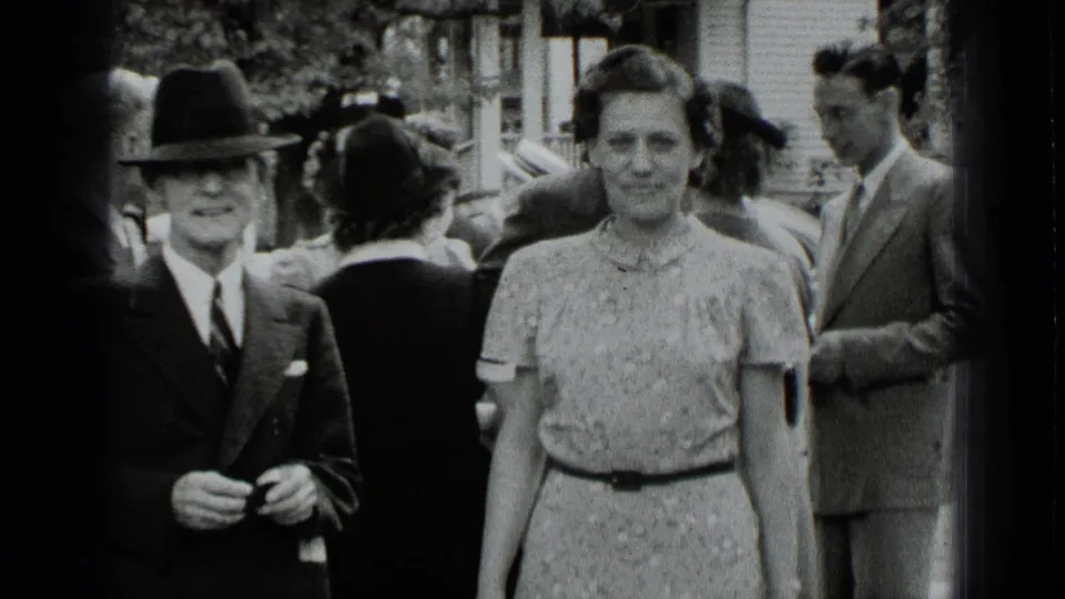 A man in a dark brimmed hat and a woman in a patterned dress face the camera during a 1939 New York outdoor gathering.