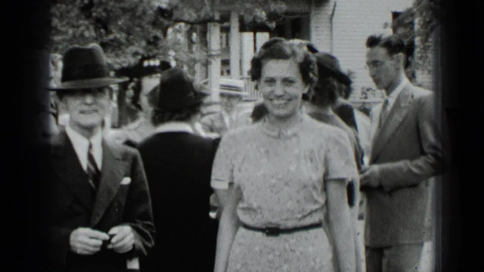 1939 New York home-movie frame of formally dressed adults at an outdoor gathering, with a suited man in a fedora and a smiling woman.