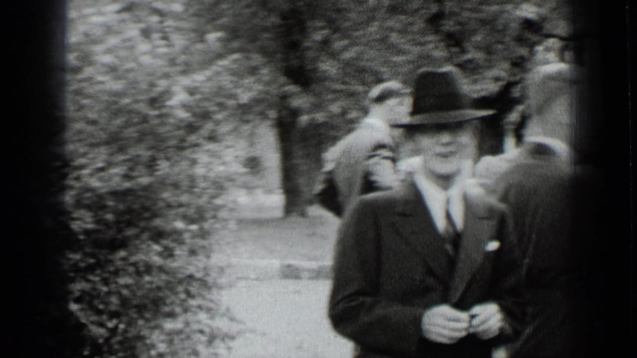 1939 New York black-and-white frame showing a suited man in a fedora near trees and a walkway at an outdoor social gathering.