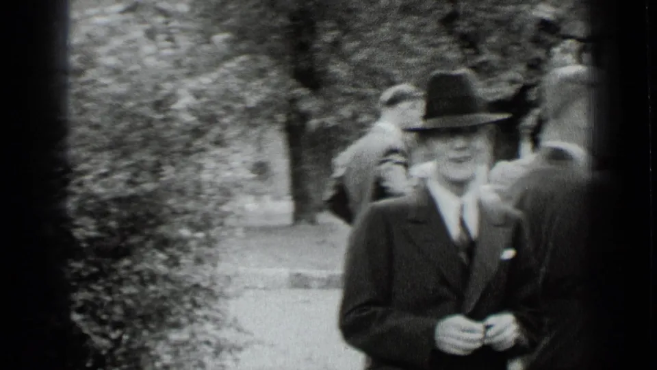1939 New York black-and-white frame showing a suited man in a fedora near trees and a walkway at an outdoor social gathering.