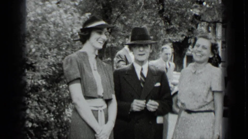 1939 New York home-movie frame of two women in dresses and hats standing with a suited man at a garden gathering.