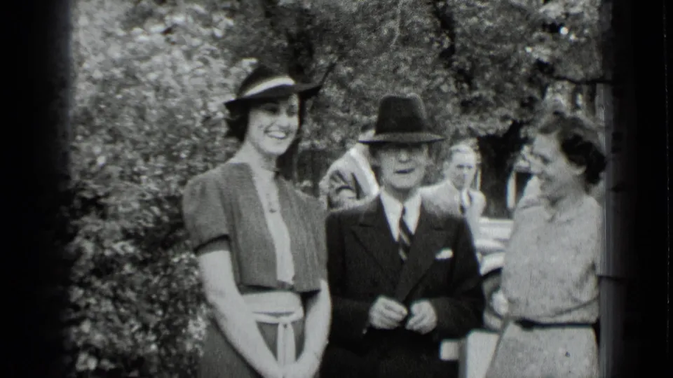 1939 New York black-and-white frame of formally dressed adults standing together outdoors, with trees and shrubbery behind them.