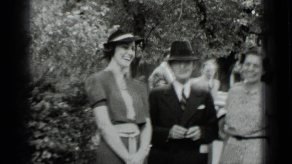 1939 New York home-movie frame showing women in dresses and hats with a suited man in a fedora outdoors near trees.