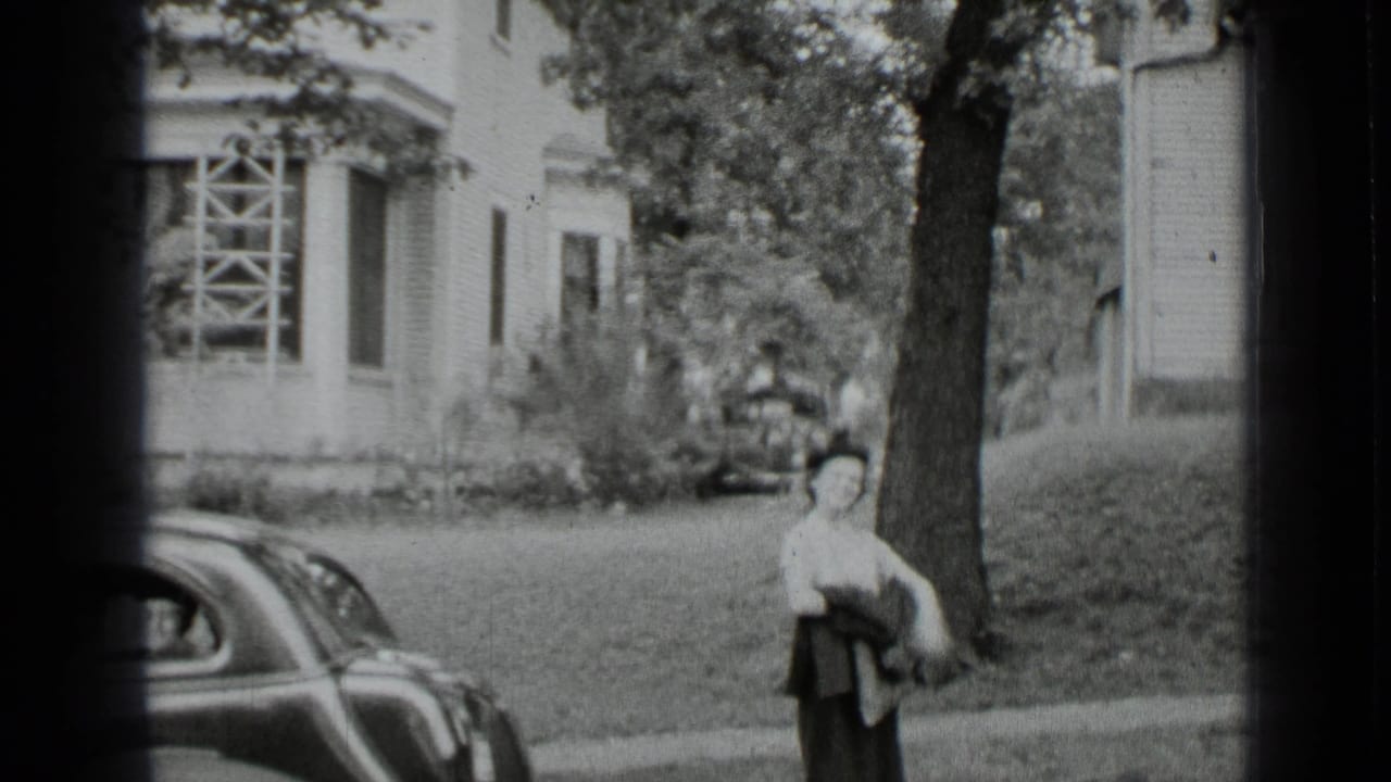 Black-and-white 1939 New York frame of a woman standing near a tree and vintage car outside a residential house.