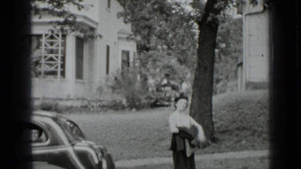 Black-and-white 1939 New York frame of a woman standing near a tree and vintage car outside a residential house.