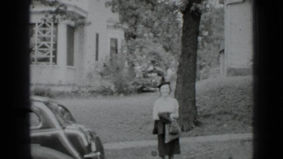 1939 New York home-movie frame showing a woman on a residential sidewalk beside a large tree and vintage car.
