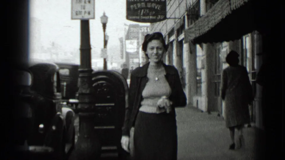 Black-and-white 1939 New York frame of a woman walking past storefronts, parked cars, and a visible perm-wave sign.