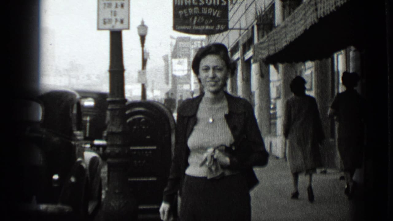 Woman walking toward the camera on a city sidewalk beside parked cars and storefronts.
