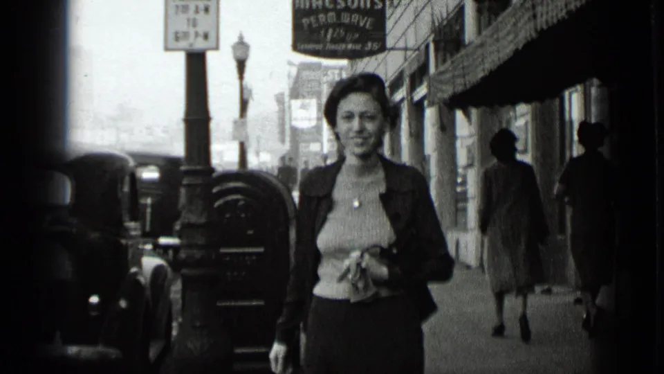 In 1939 New York, a woman walks toward the camera on a city sidewalk with parked cars, pedestrians, and storefront signs nearby.