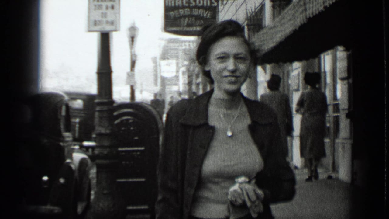 Smiling woman closer to the camera with storefronts, pedestrians, and parked cars behind her.