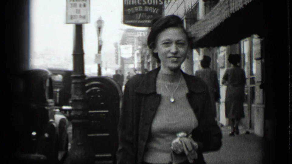 A smiling woman in 1939 New York walks close to the camera, with parked cars, pedestrians, and sidewalk storefronts in the background.