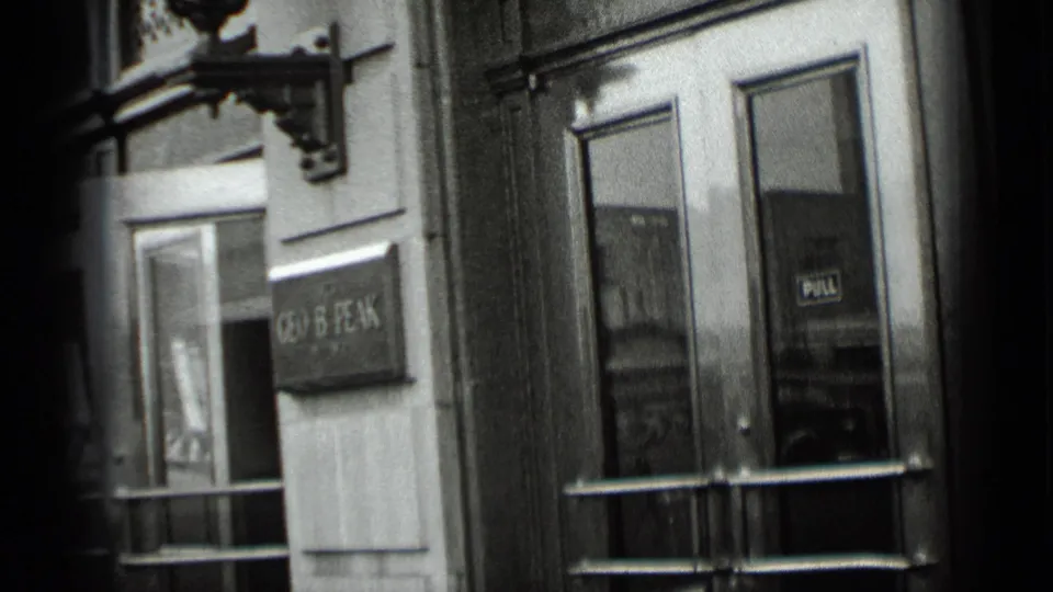 A handheld 1939 New York street view shows storefront windows, a doorway, building facade, and a blurred sign in soft black-and-white focus.