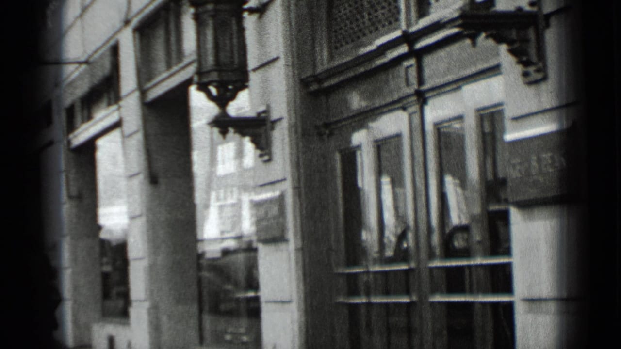 1939 New York sidewalk frame with a concrete or stone facade, glass doors, window reflections, and a decorative sconce above the entrance.