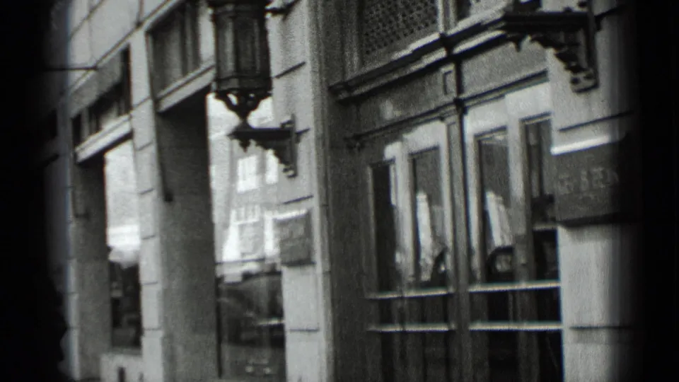 1939 New York sidewalk frame with a concrete or stone facade, glass doors, window reflections, and a decorative sconce above the entrance.