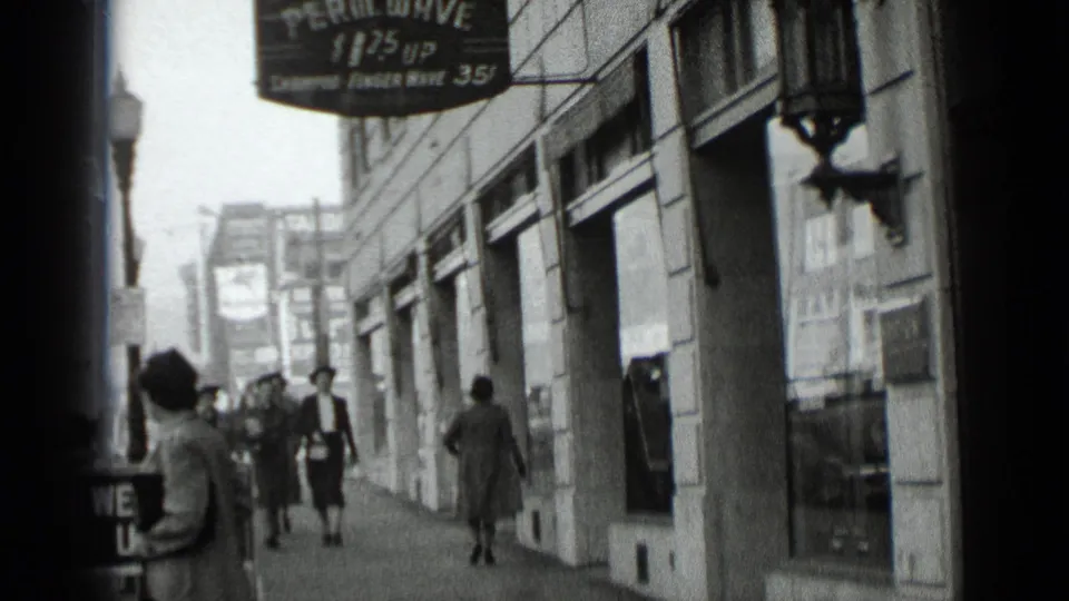1939 New York home-movie frame showing pedestrians on a city sidewalk beneath a hanging sign advertising a film wave service.