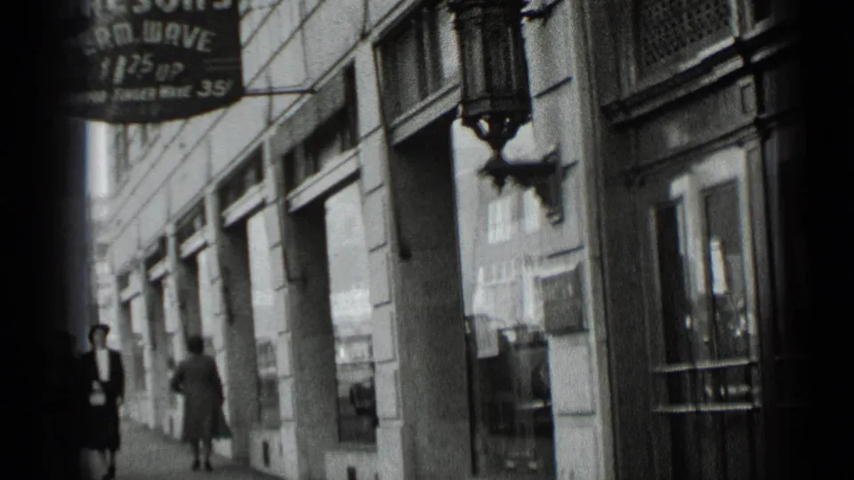 1939 New York 8mm frame with pedestrians passing a commercial building facade, glass windows, a wall sconce, and a partially readable hanging sign.