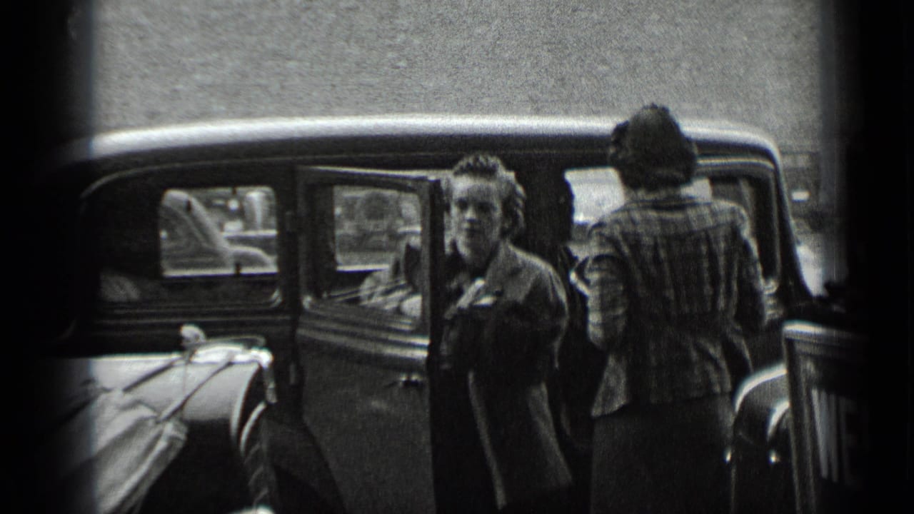 Two women stand beside a parked vintage car on a city street.