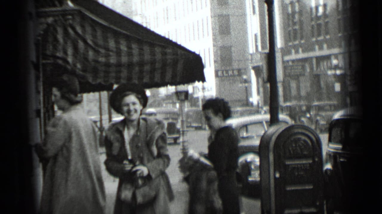 Pedestrians stand near a storefront awning with parked cars and a parking meter on the street.
