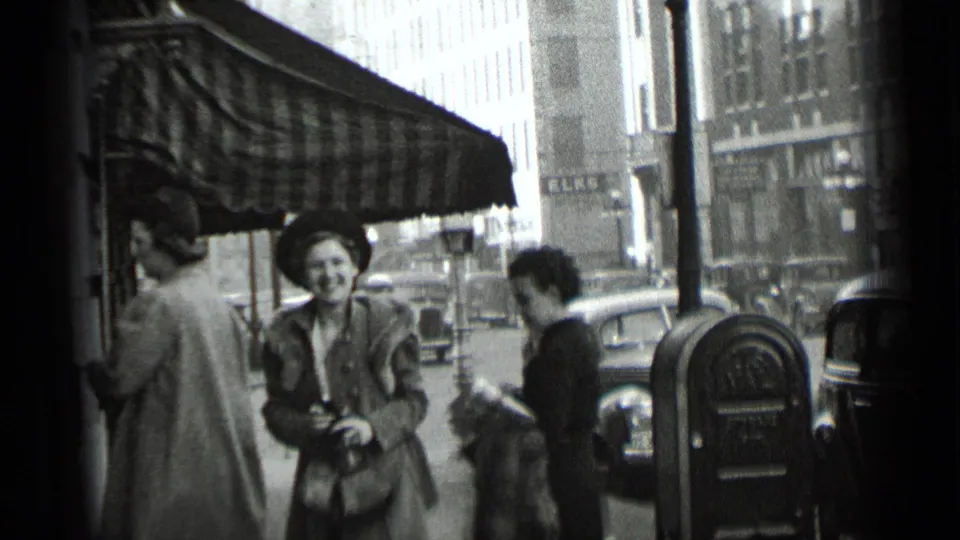 1939 New York street view with pedestrians in coats and hats near an awning, parked cars, storefronts, and a parking meter.