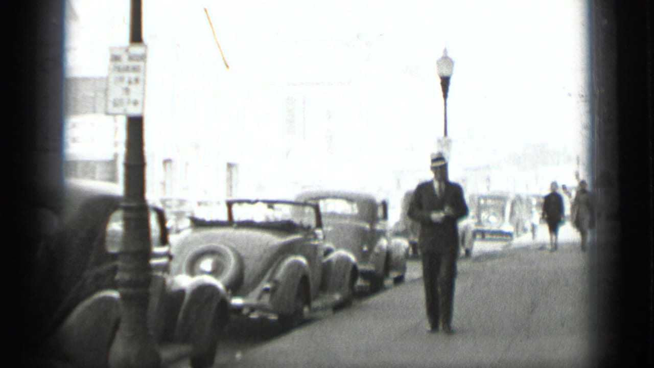 A suited man walks along a sidewalk past parked cars and a street lamp.