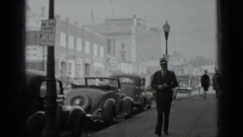 Black-and-white 8mm frame of a suited man on a New York sidewalk, with curbside cars and distant pedestrians behind him.