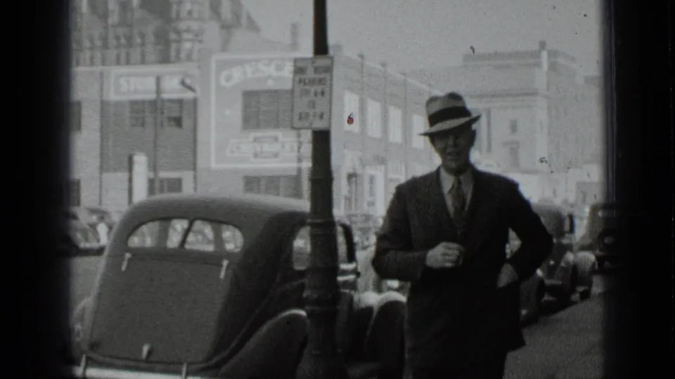 Close street view of a suited man in a fedora passing the camera on a 1939 New York sidewalk, with cars and storefront signs behind.