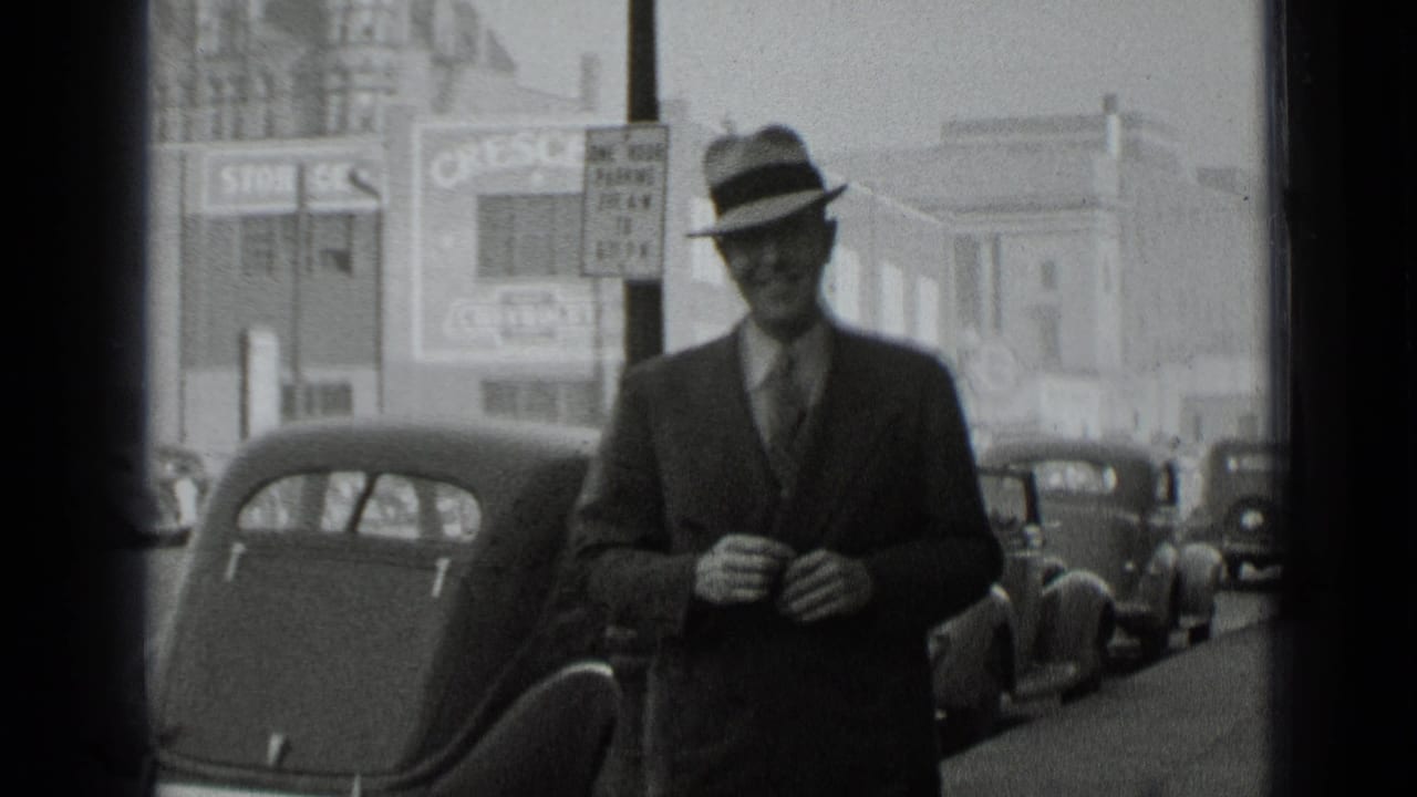 Man in a fedora walks on a city street beside parked 1930s cars.