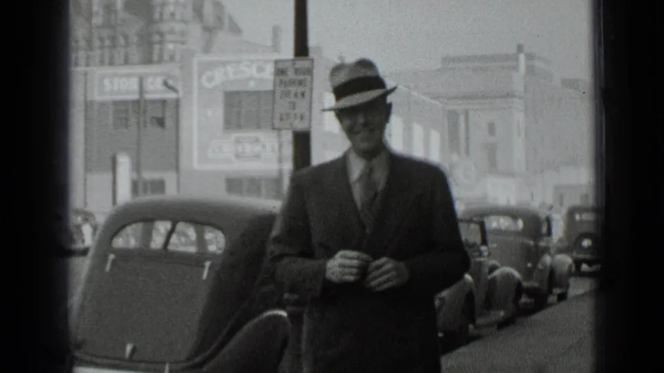 In 1939 New York footage, a suited man in a fedora walks toward the camera near parked classic cars and storefront signs.